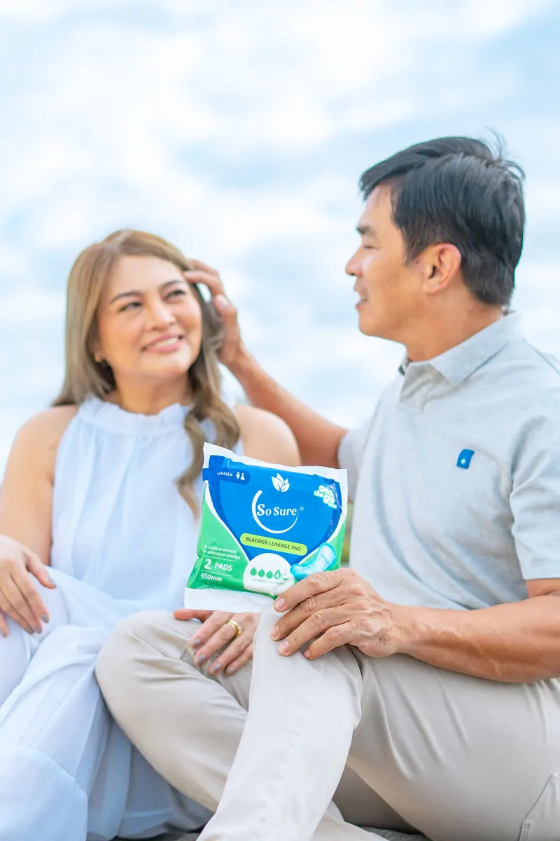 An older Filipino couple sit together against a bright sky as the man holds a So Sure pack.