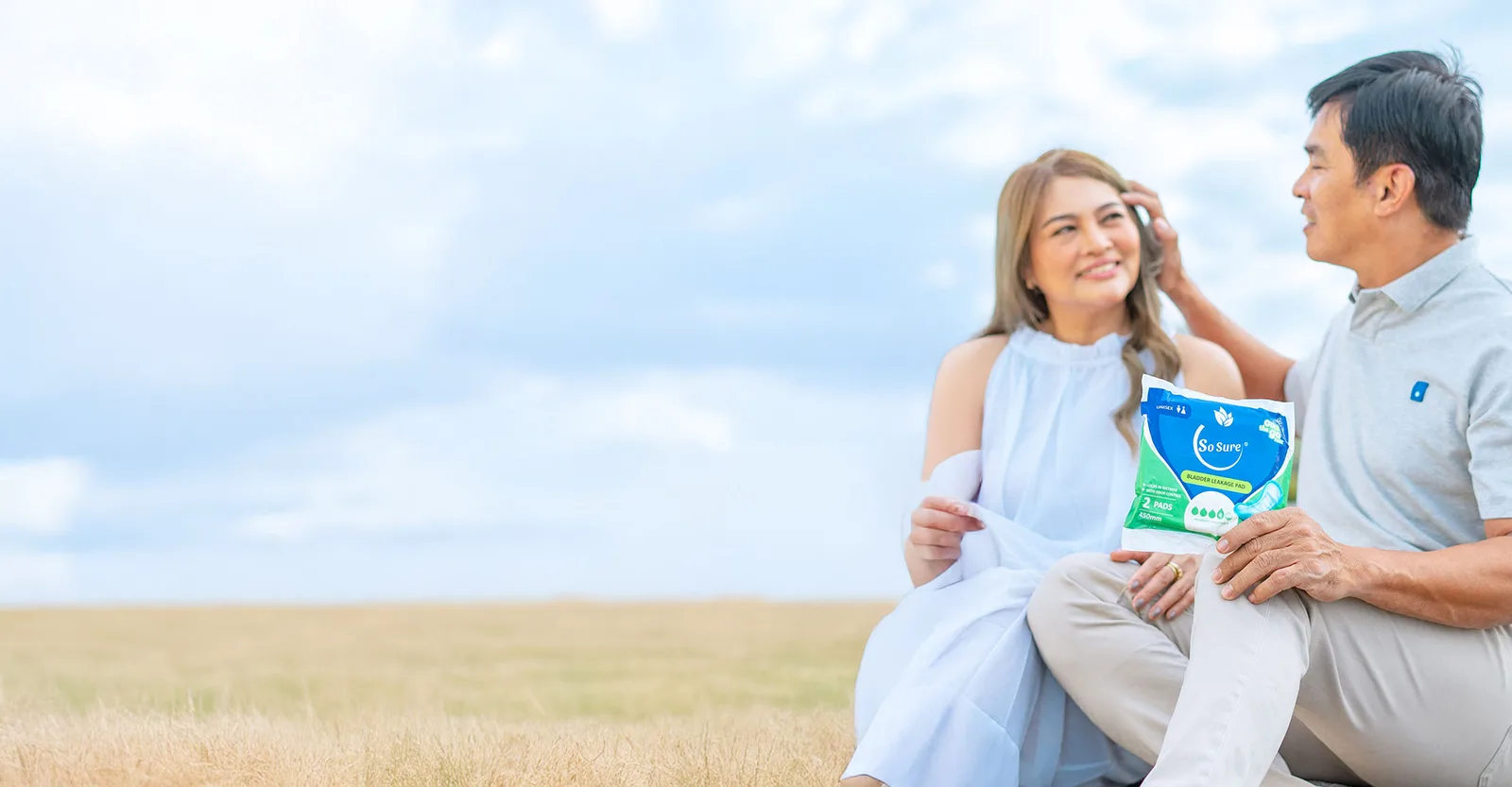 A smiling older Filipino couple sit close together against a bright sky as the woman holds a So Sure pack.